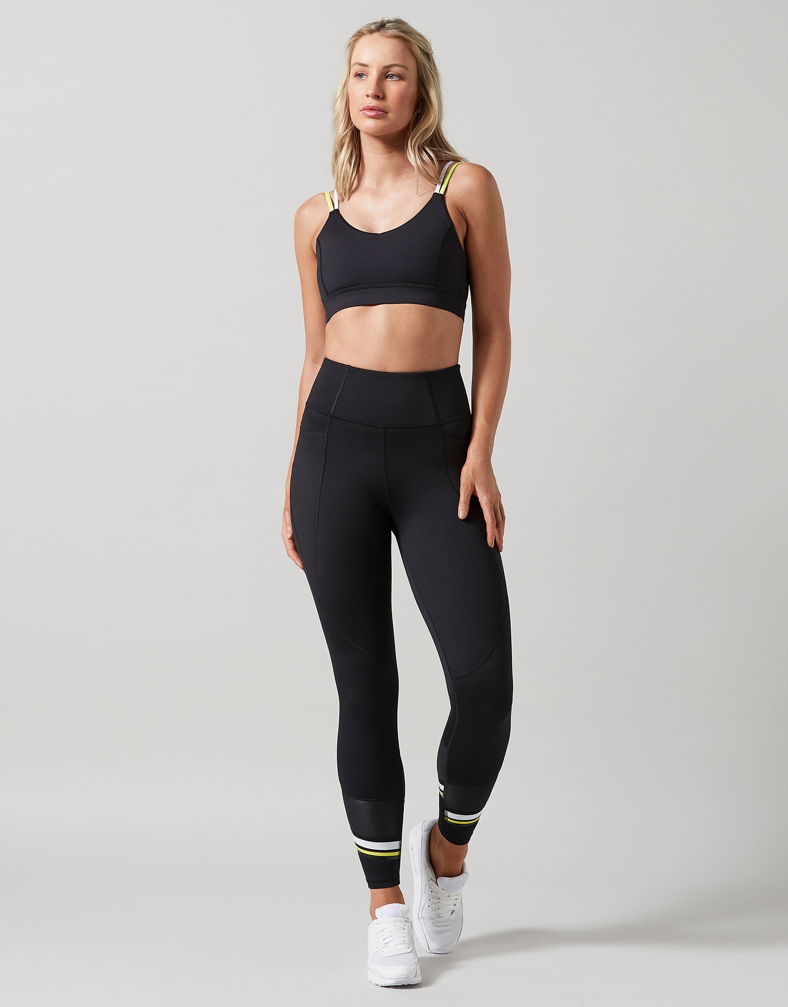 Woman wearing a black sports bra and leggings with white sneakers on a light gray background