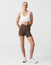 Woman wearing a white sports bra and brown shorts on a light gray background
