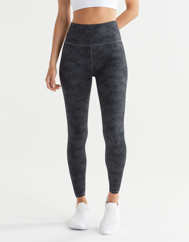 A model wearing high-waisted full-length black stucco leggings and a sports bra, with white sneakers.