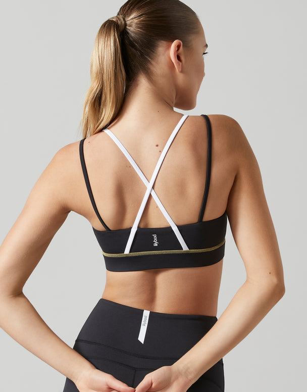Rear view of a woman wearing a black sports bra with white crisscross back straps and a thin gold accent band, paired with black leggings, against a light gray studio background.