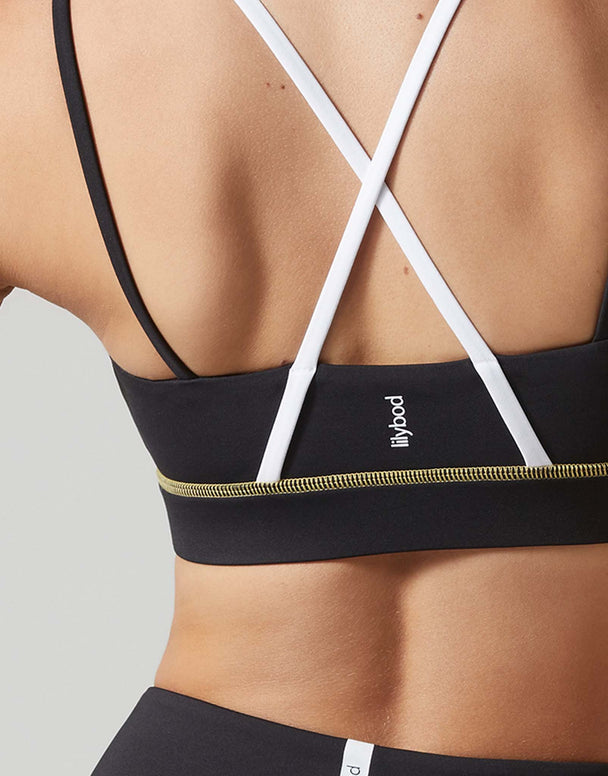 Close-up back view of a person wearing a black sports bra with white crisscross straps and a gold zipper detail along the underband. The brand name 'lilypod' is printed in white on the center back.