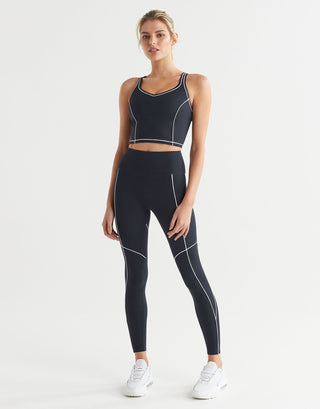 A model wearing a sports bra and full-length navy leggings with white contrast seaming and a V-point waistband, paired with white sneakers.