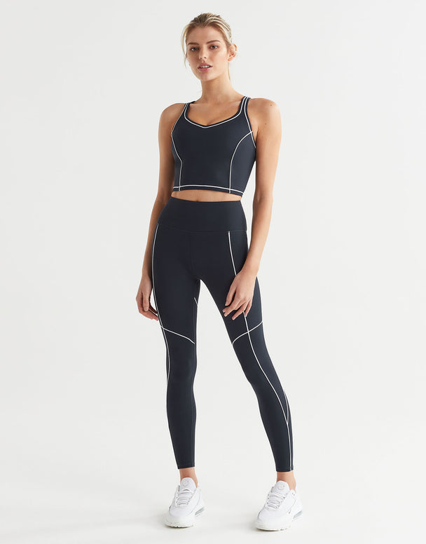 A model wearing a sports bra and full-length navy leggings with white contrast seaming and a V-point waistband, paired with white sneakers.