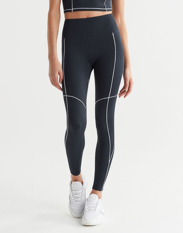 A person wearing full-length navy leggings with white contrast seaming and a V-point waistband, paired with white sneakers.