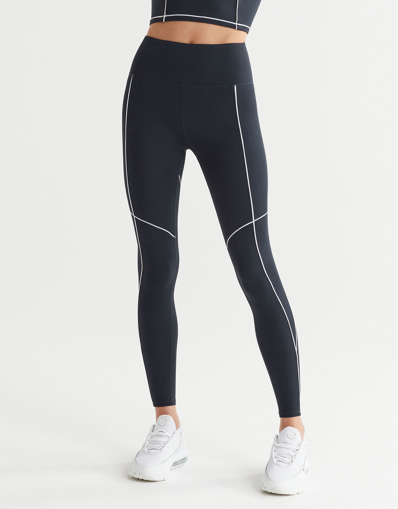 A woman wearing full-length navy leggings with white contrast seaming and a V-point waistband, paired with white sneakers.