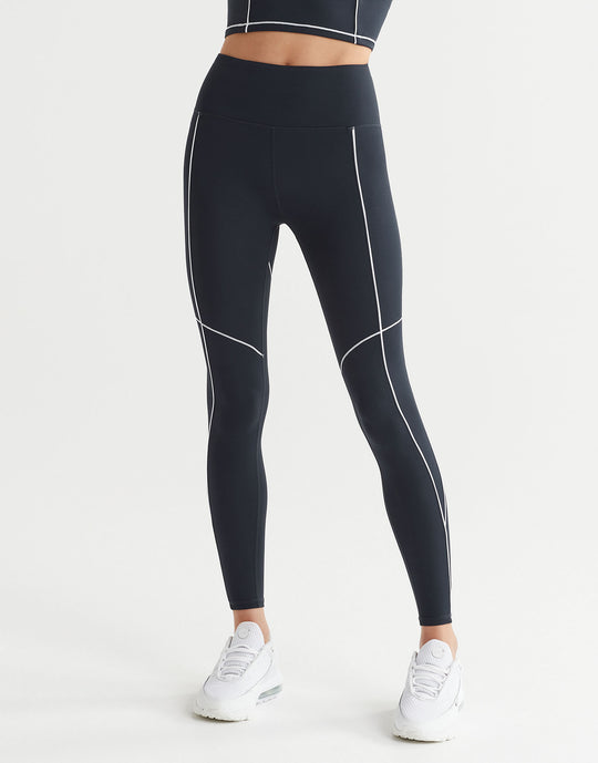 A woman wearing full-length navy leggings with white contrast seaming and a V-point waistband, paired with white sneakers.