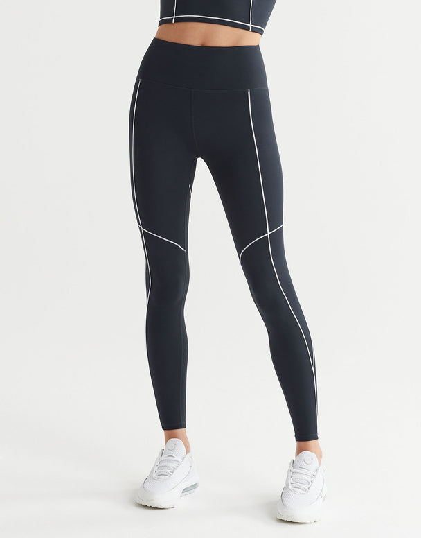 A woman wearing full-length navy leggings with white contrast seaming and a V-point waistband, paired with white sneakers.