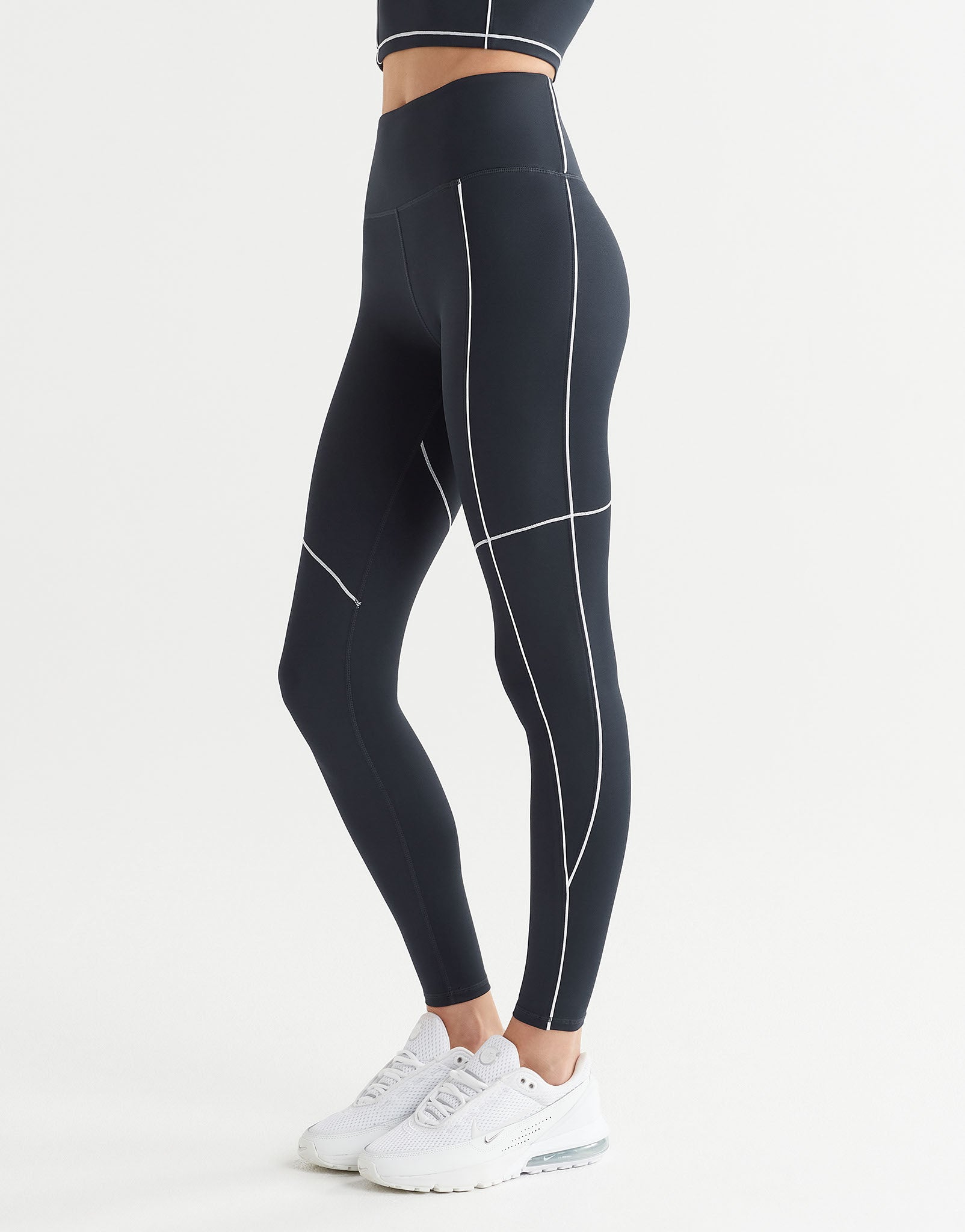 Black high-waisted athletic leggings with white contrast stitching, shown on a model from the waist down wearing white sneakers against a light gray studio background.