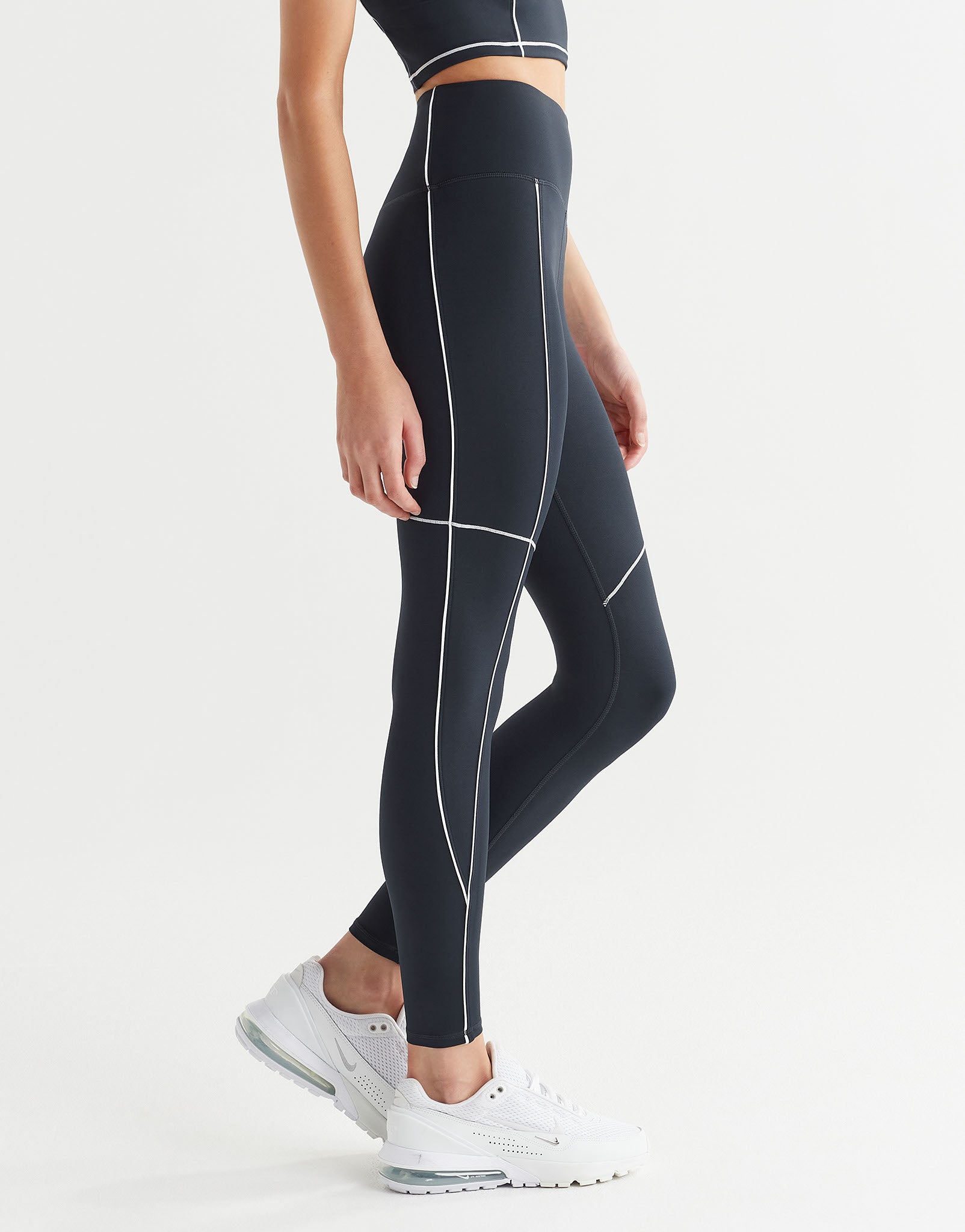 A person wearing full-length navy leggings with white contrast seaming and a V-point waistband, paired with white sneakers and a sports bra.
