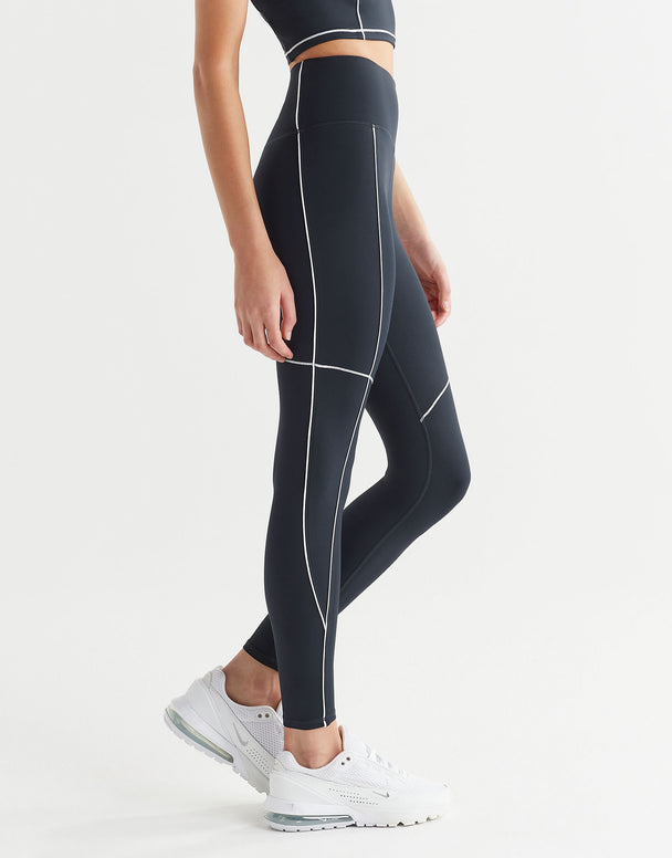 A person wearing full-length navy leggings with white contrast seaming and a V-point waistband, paired with white sneakers and a sports bra.