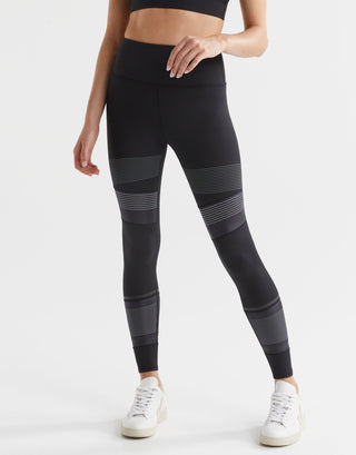 A model wearing high-waisted black leggings with micro dot/stripe print accents, paired with a black sports top and white sneakers.