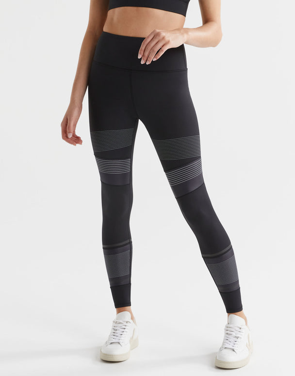 A model wearing high-waisted black leggings with micro dot/stripe print accents, paired with a black sports top and white sneakers.