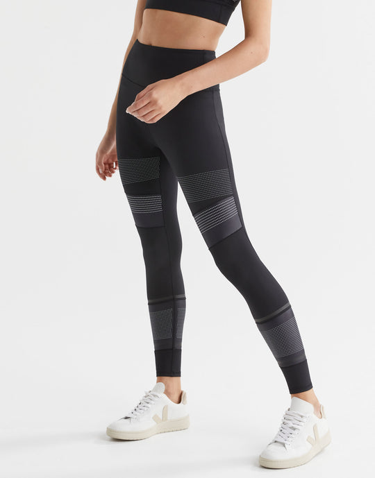 A woman wearing high-waisted black leggings with micro dot/stripe print accents, paired with a black sports top and white sneakers.