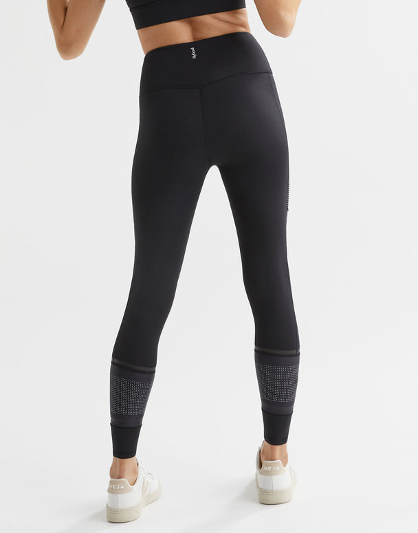A model wearing high-waisted black leggings with micro dot/stripe print accents, paired with a black sports top and white sneakers. Rear View.