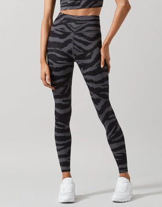 A model wearing high-waisted leggings with a dark gray and black zebra stripe pattern, paired with white sneakers, shown from a rear three-quarter angle against a light gray studio background.