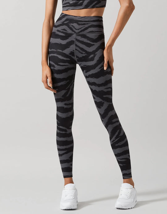 A model wearing high-waisted leggings with a dark gray and black zebra stripe pattern, paired with white sneakers, shown from a rear three-quarter angle against a light gray studio background.