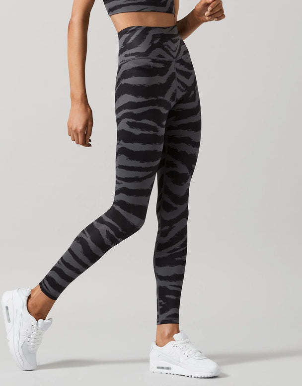 A model wearing high-waisted leggings with a dark gray and black tiger stripe pattern, paired with white sneakers, shown from the waist down on a light gray studio background.