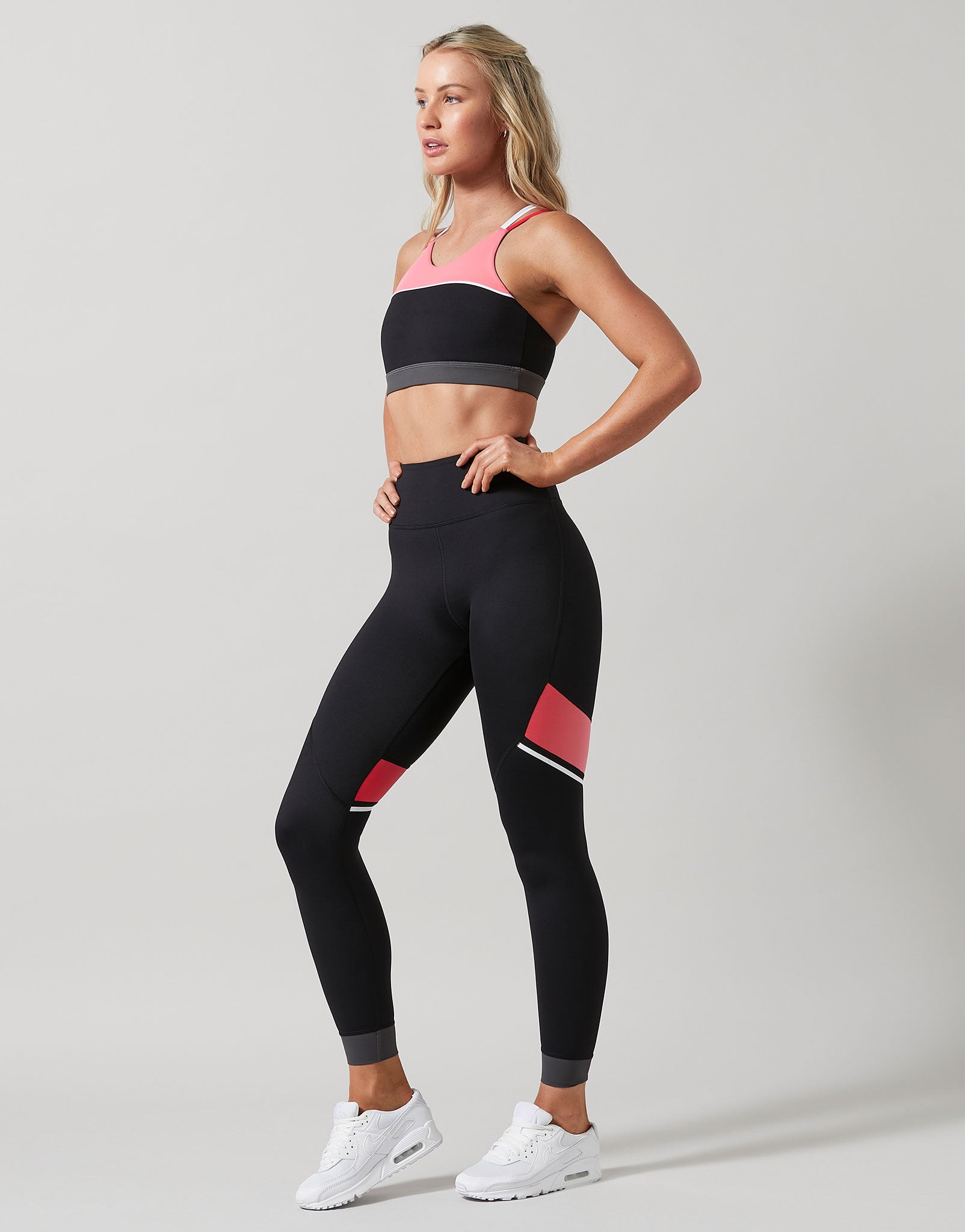 A woman wearing black leggings and a sports bra with pink and grey accents on a grey background.
