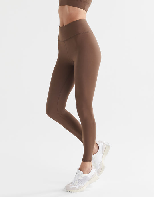 A woman wearing a pair of full-length brown leggings with a high-waisted fit, paired with a matching sports bra and white sneakers.