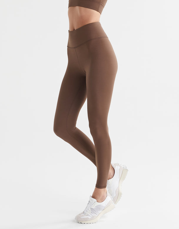 A woman wearing a pair of full-length brown leggings with a high-waisted fit, paired with a matching sports bra and white sneakers.