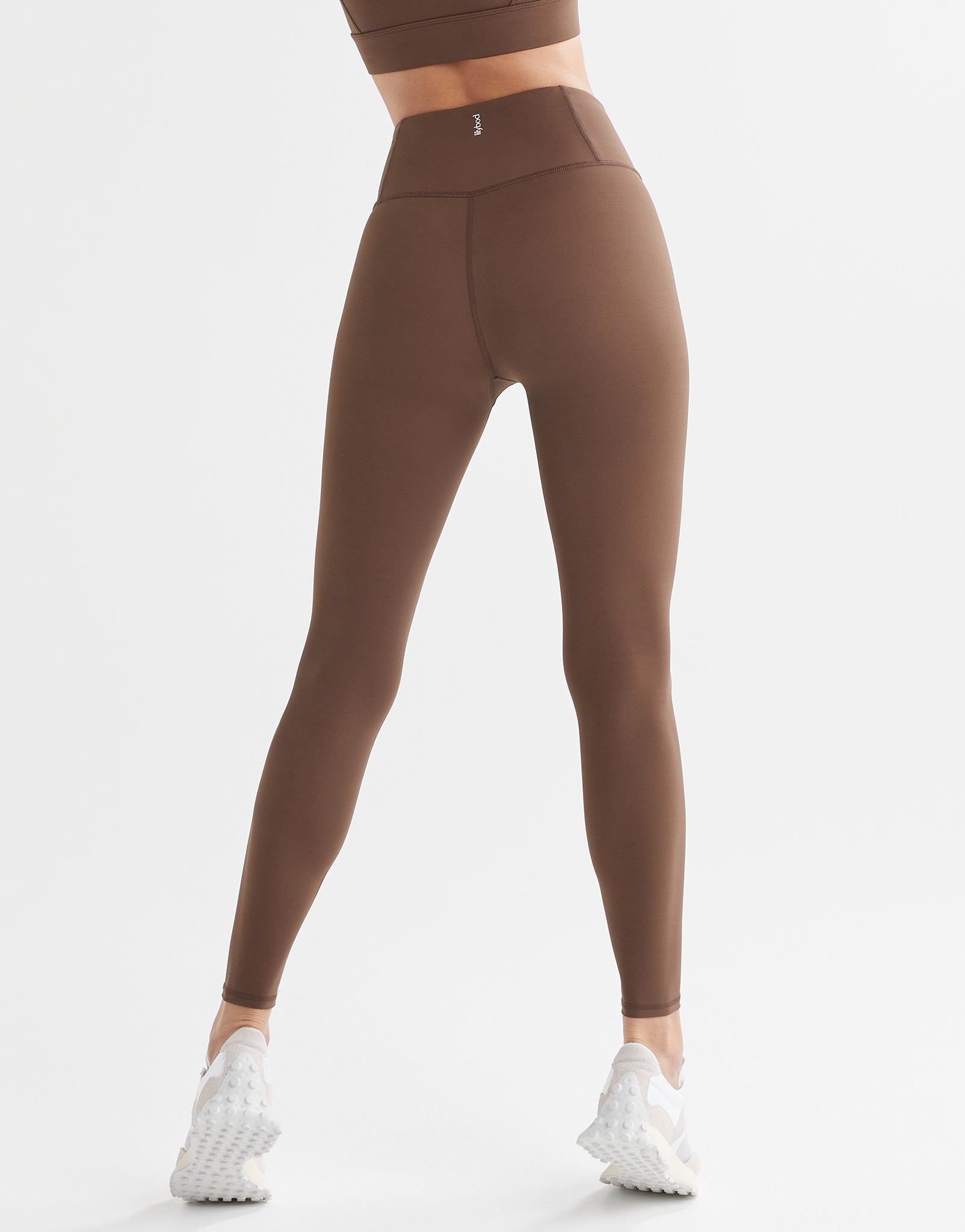 A model in a pair of full-length brown leggings with a high-waisted fit, paired with a matching sports bra and white sneakers.