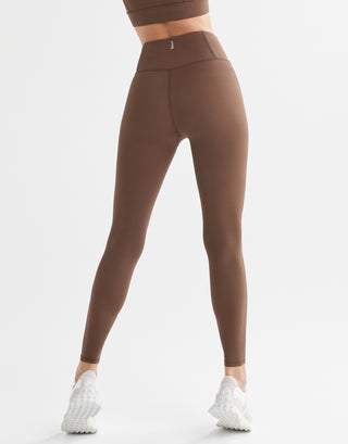 A model in a pair of full-length brown leggings with a high-waisted fit, paired with a matching sports bra and white sneakers.