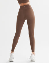 A model in a pair of full-length brown leggings with a high-waisted fit, paired with a matching sports bra and white sneakers.