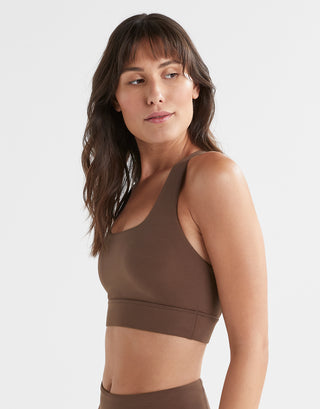 A woman wearing a brown Lilybod Astrid sports bra with a scoop front neckline and wider straps and brown leggings.