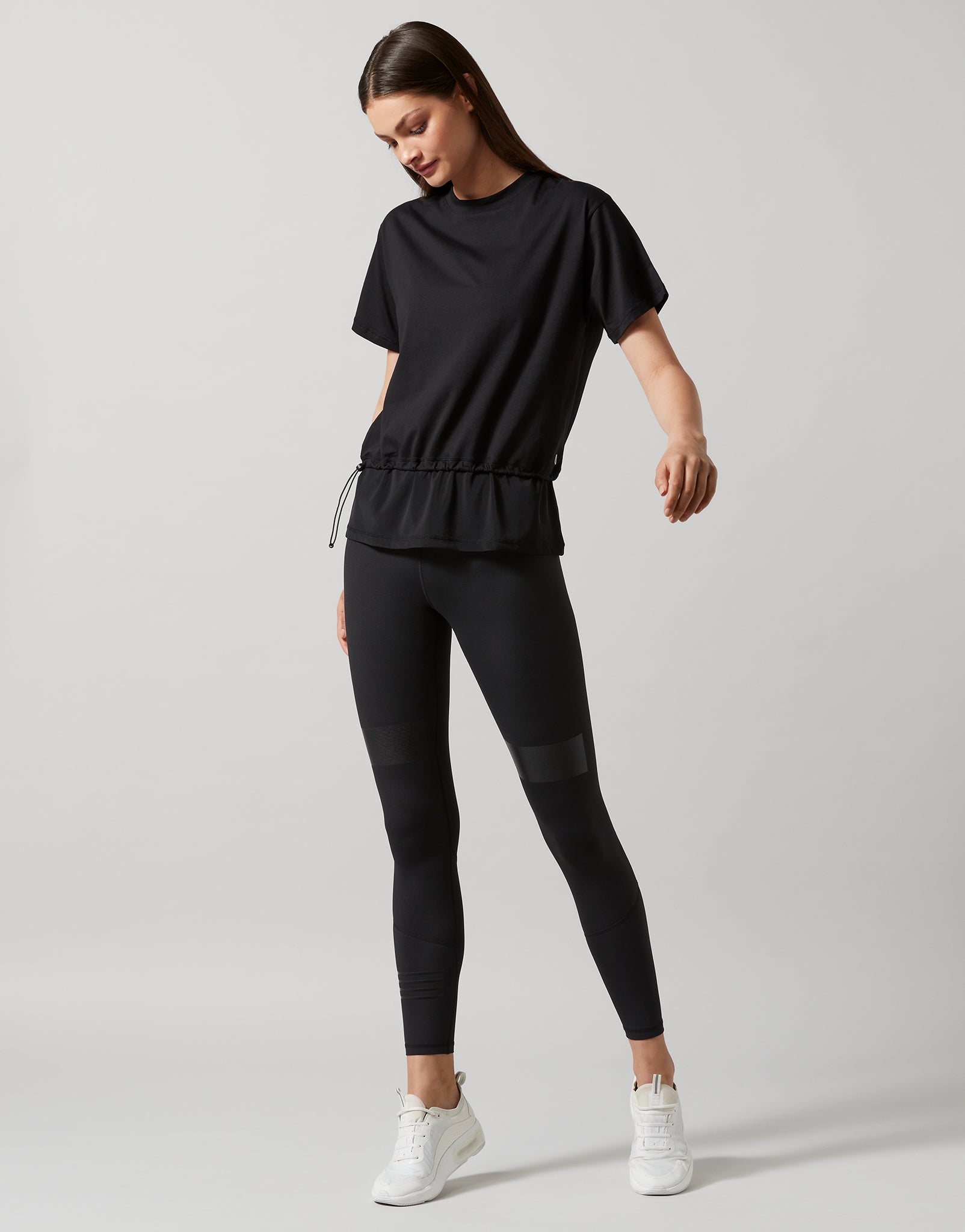 A woman wearing a black, oversized, short-sleeved t-shirt with accent stripes and hardware on a model wearing black pants and white runners.