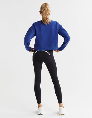 A woman wearing a blue cropped fleece crew sweater with black leggings. The sweater has a crew neck and the brand logo is visible. Rear view.