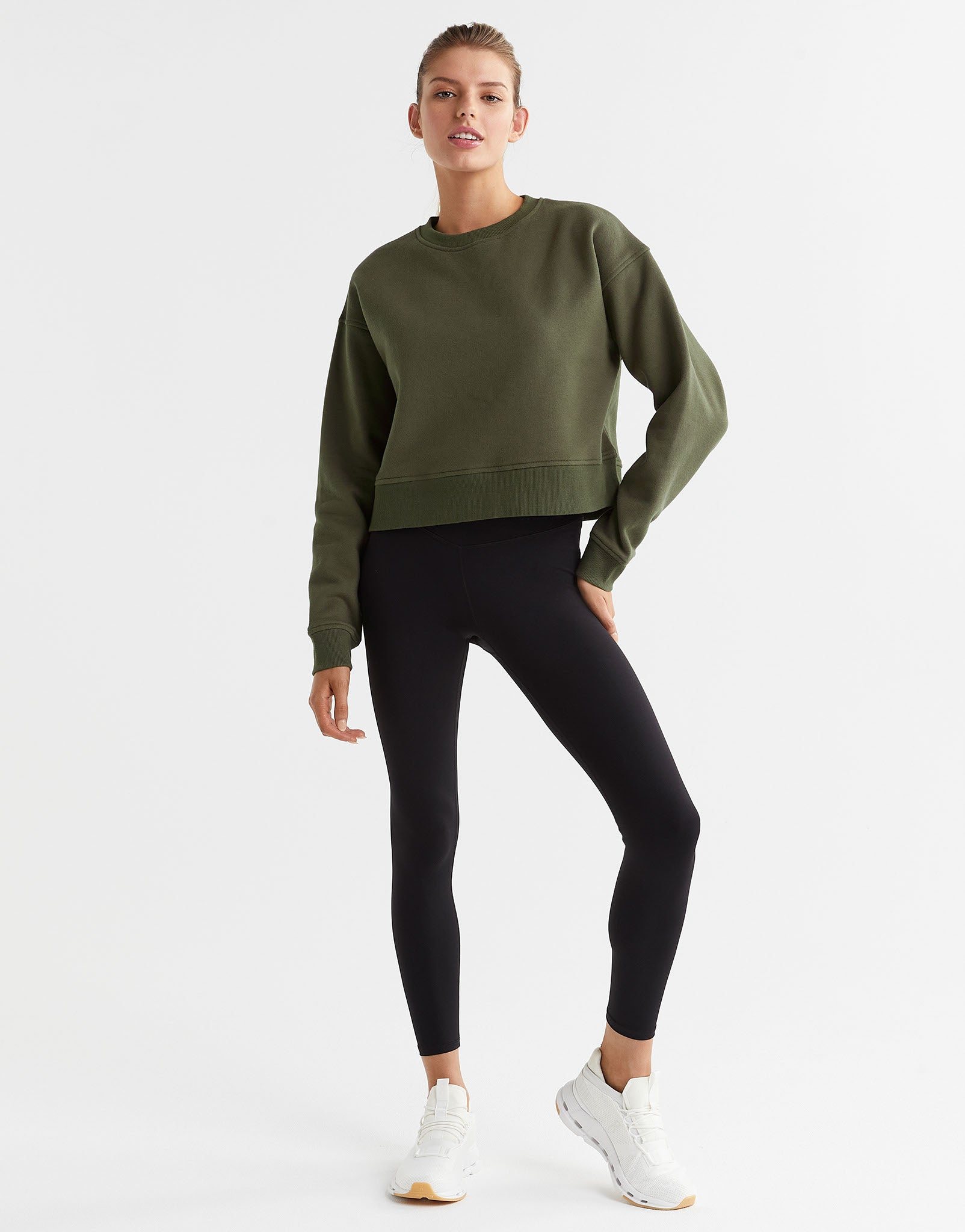A woman wearing an olive green sweater and black leggings and white sneakers.