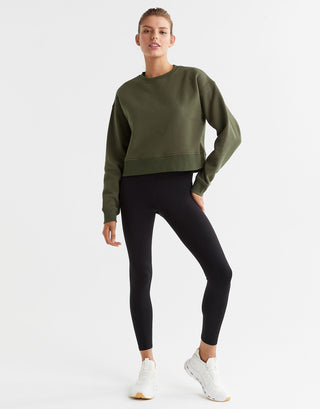 A woman wearing an olive green sweater and black leggings and white sneakers.