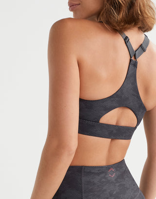 Rear view of a woman wearing a dark gray patterned sports bra with cutout back details and matching high-waisted leggings against a light gray studio background.