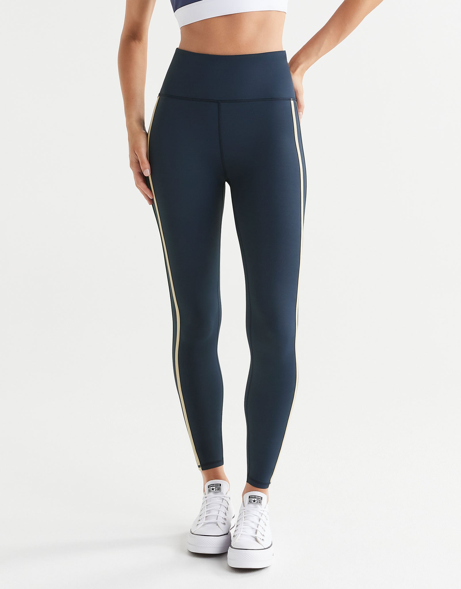 A woman wearing navy blue leggings with white runners on a plain background.
