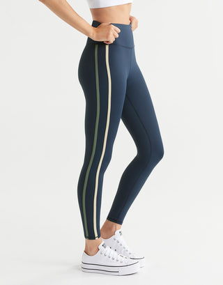 A woman wearing navy blue leggings with stripes and white runners on a plain background.