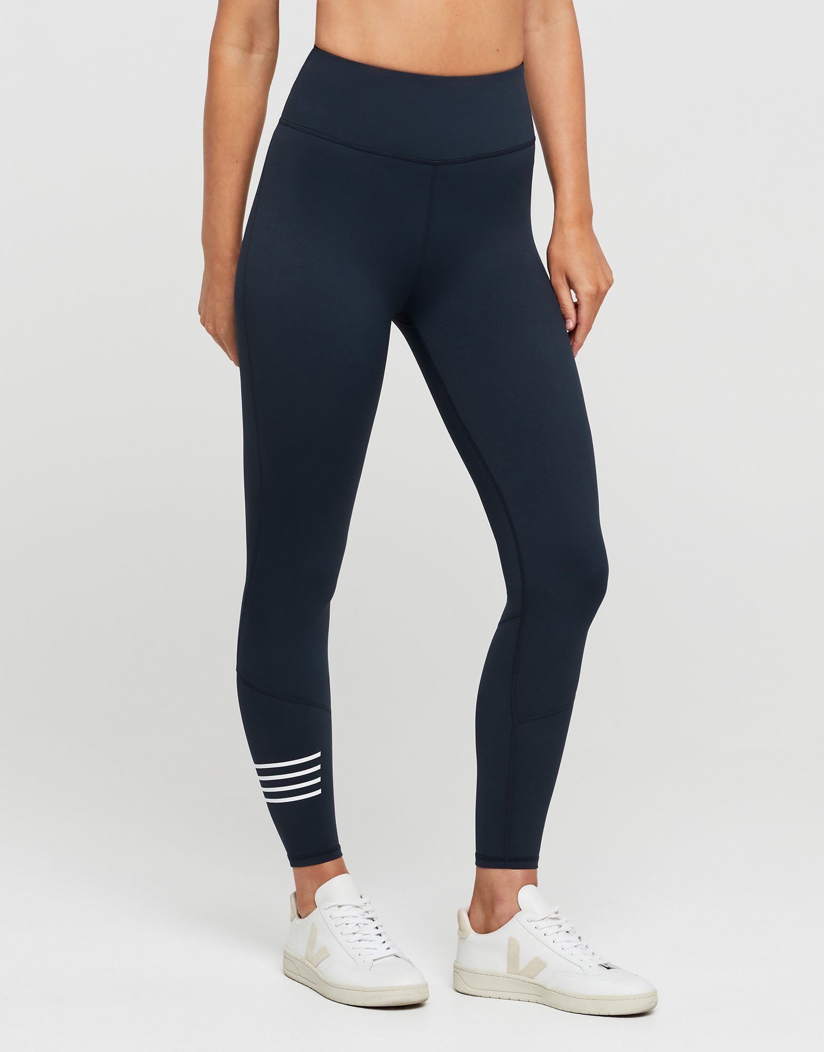 A model wearing high-waisted navy blue leggings with three white horizontal stripes near the ankle, paired with white sneakers, shown from the waist down on a light gray studio background.
