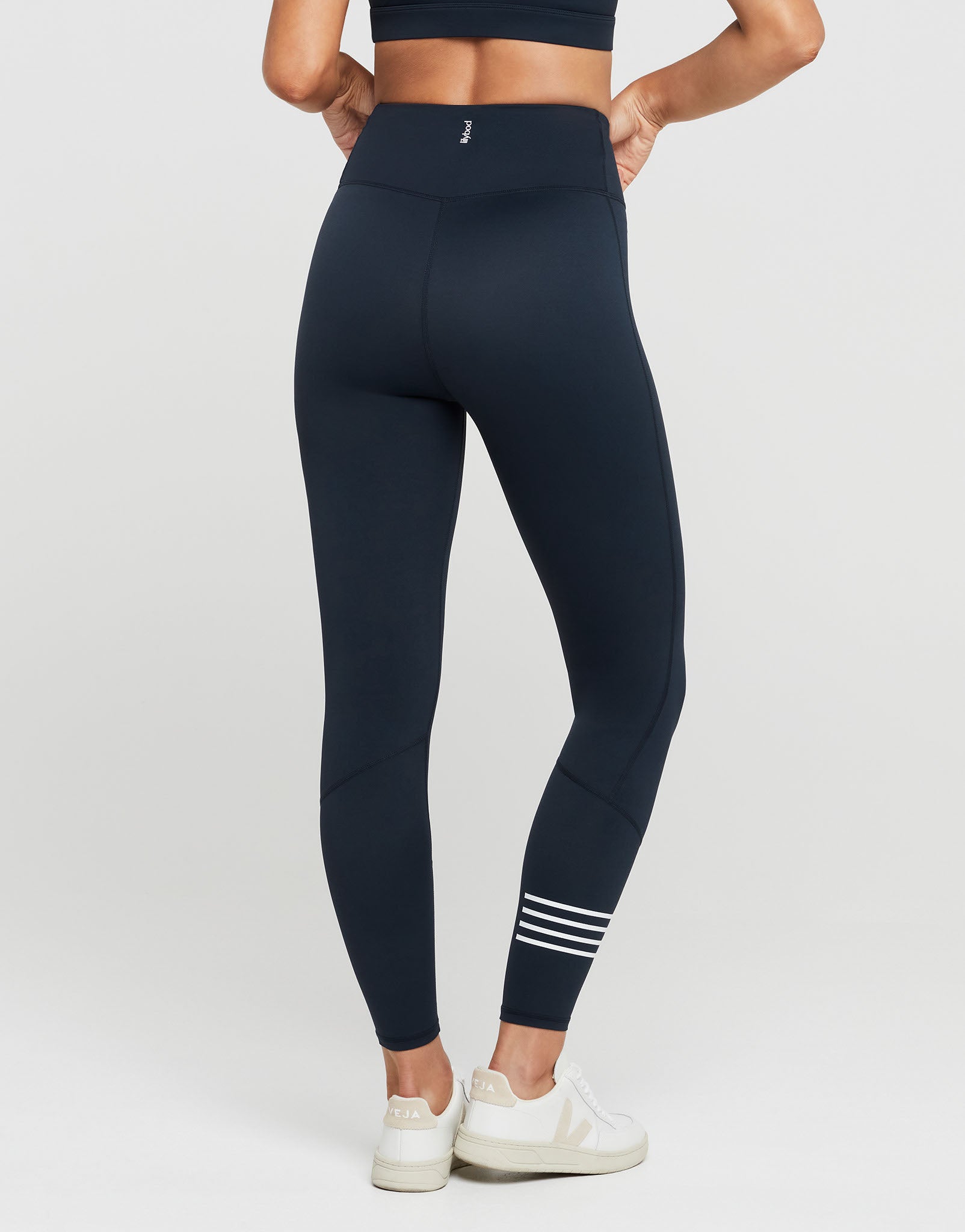 A model from the back wearing high-waisted navy blue leggings with white stripes on the lower calf, paired with white sneakers, against a light gray studio background.
