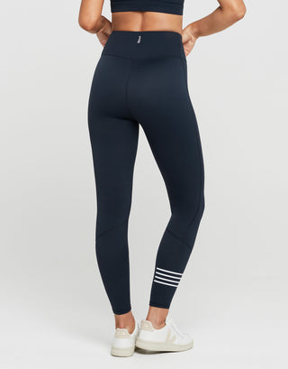 A model from the back wearing high-waisted navy blue leggings with white stripes on the lower calf, paired with white sneakers, against a light gray studio background.