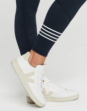 White sneakers with a brand logo worn with navy leggings featuring white stripes on a light gray background