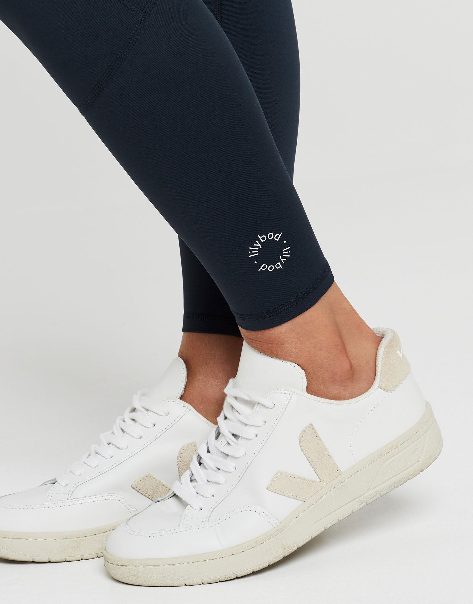White Veja low-top sneakers with beige side stripes and white laces, shown on a model wearing navy blue leggings against a light gray studio background.