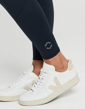 Close up of a woman wearing navy leggings with a logo and white sneakers on a light gray background