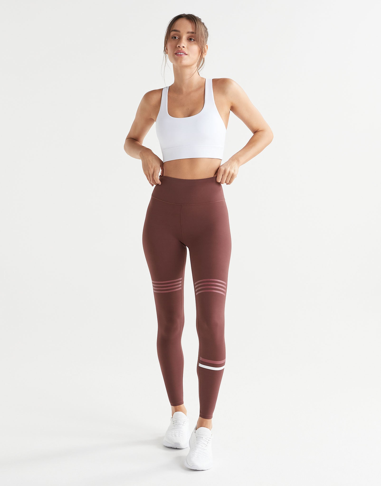 A woman modeling high-waisted athletic leggings in a dusty rose-brown color with white stripes near the knees and ankles, paired with a white sports bra and white sneakers, against a plain white studio background.