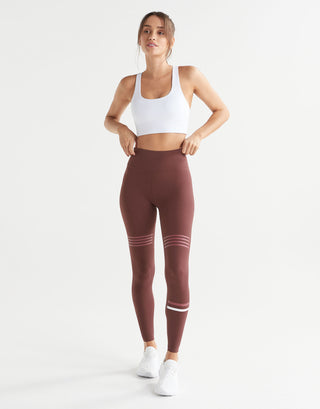 A woman modeling high-waisted athletic leggings in a dusty rose-brown color with white stripes near the knees and ankles, paired with a white sports bra and white sneakers, against a plain white studio background.