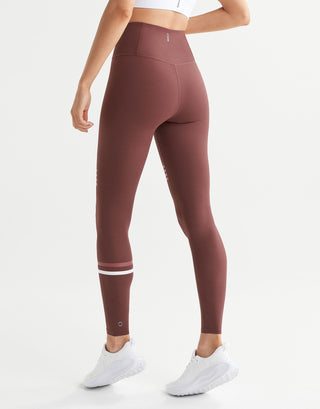 A model from the back wearing high-waisted athletic leggings in a dusty rose-brown color with white stripes near the ankles, paired with white sneakers on a light gray studio background.