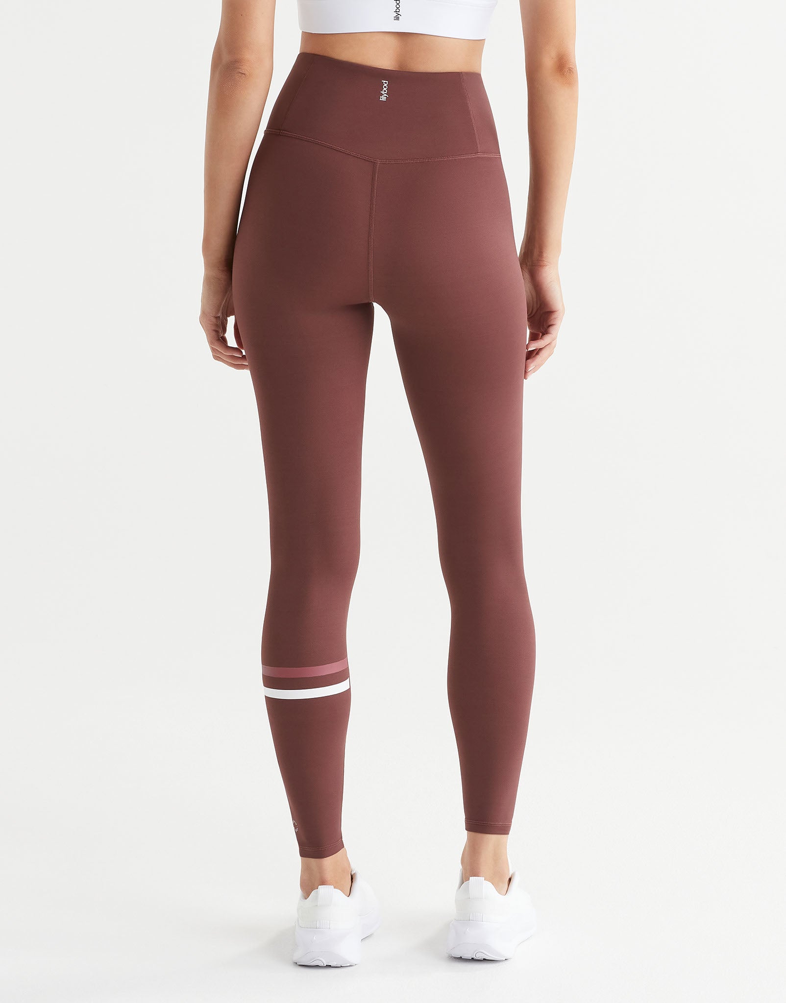 A model from the back wearing high-waisted, form-fitting leggings in a dusty rose-brown color, featuring a double white stripe detail near the lower calf, paired with white sneakers against a clean white background.