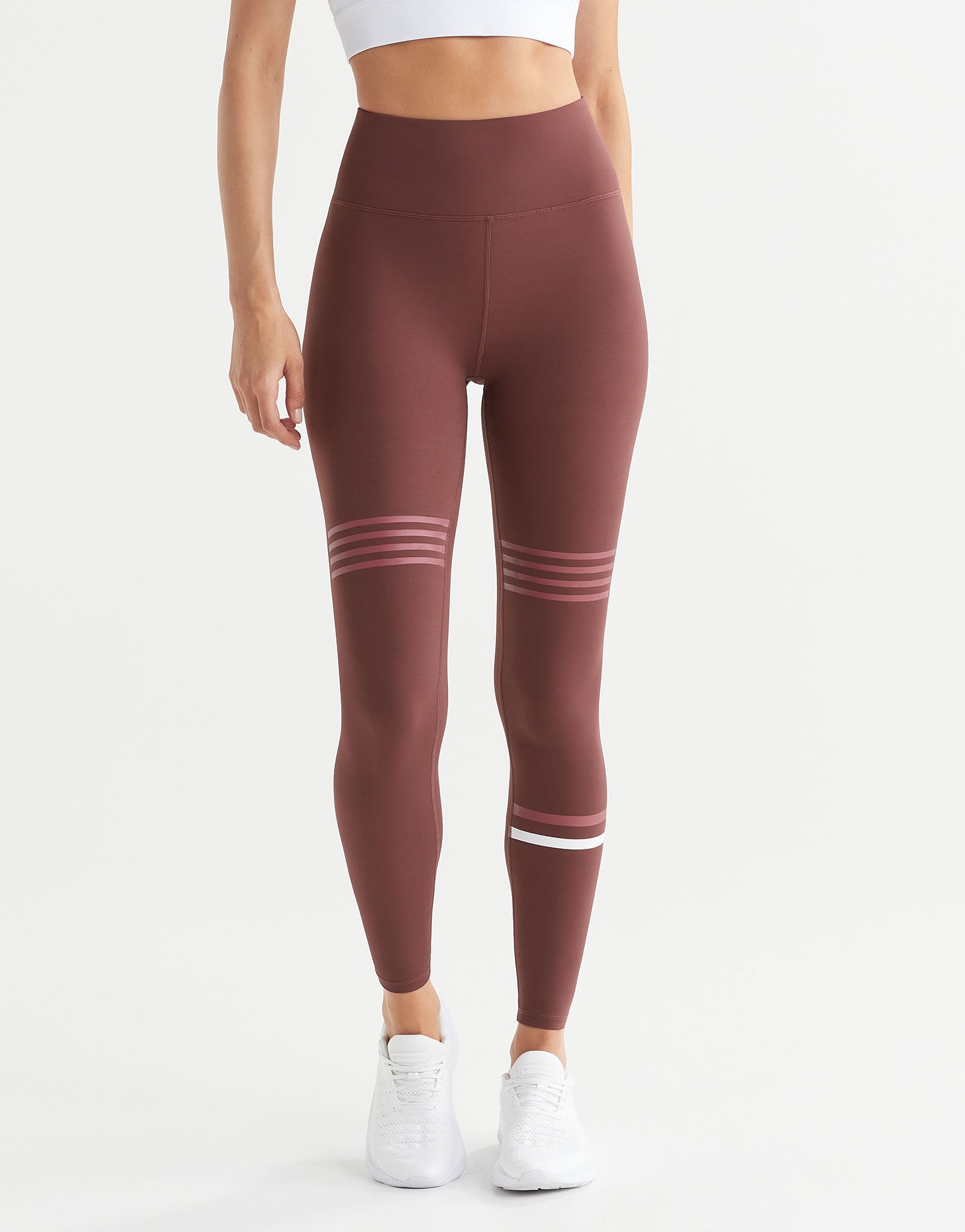 A model wearing high-waisted athletic leggings in a dusty rose-brown color, featuring three horizontal white stripes on the upper thighs and one on the lower leg, paired with white sneakers against a plain white studio background.