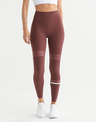 A model wearing high-waisted athletic leggings in a dusty rose-brown color, featuring three horizontal white stripes on the upper thighs and one on the lower leg, paired with white sneakers against a plain white studio background.