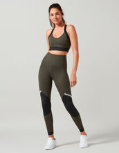 Woman wearing a black sports bra and olive green leggings on a light gray background