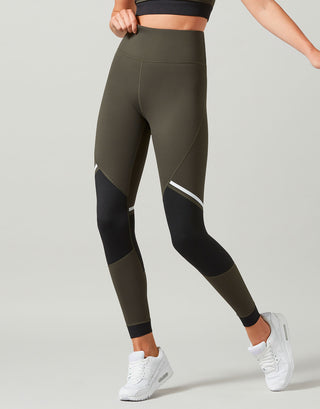 A model wearing high-waisted athletic leggings in olive green and black with white contrast panels, paired with white sneakers, against a light gray studio background.