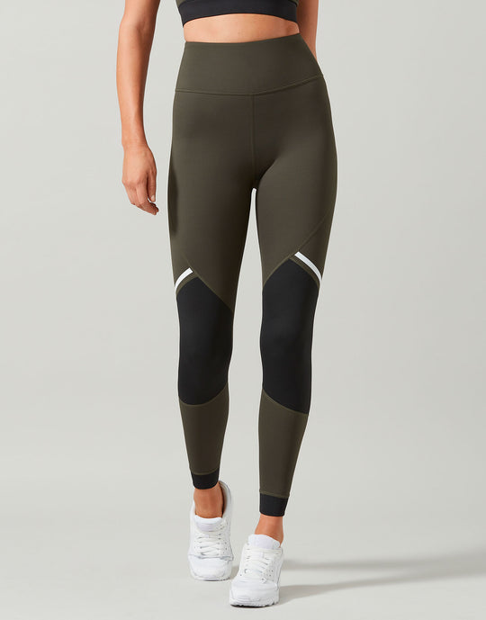 A model wearing high-waisted athletic leggings in olive green and black with white contrast panels, paired with white sneakers, against a light gray studio background.
